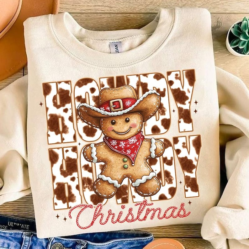 May include: A beige sweatshirt with a brown and white cow print design. The text "Howdy Hoody Christmas" is printed on the sweatshirt. A gingerbread man wearing a cowboy hat is in the center of the design.