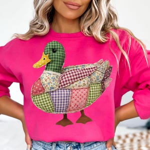May include: A hot pink sweatshirt featuring a patchwork duck design. The duck has a green checkered head, a yellow beak, and a body made of various patterned fabrics, including floral, polka dot, and gingham. The duck's feet are brown.