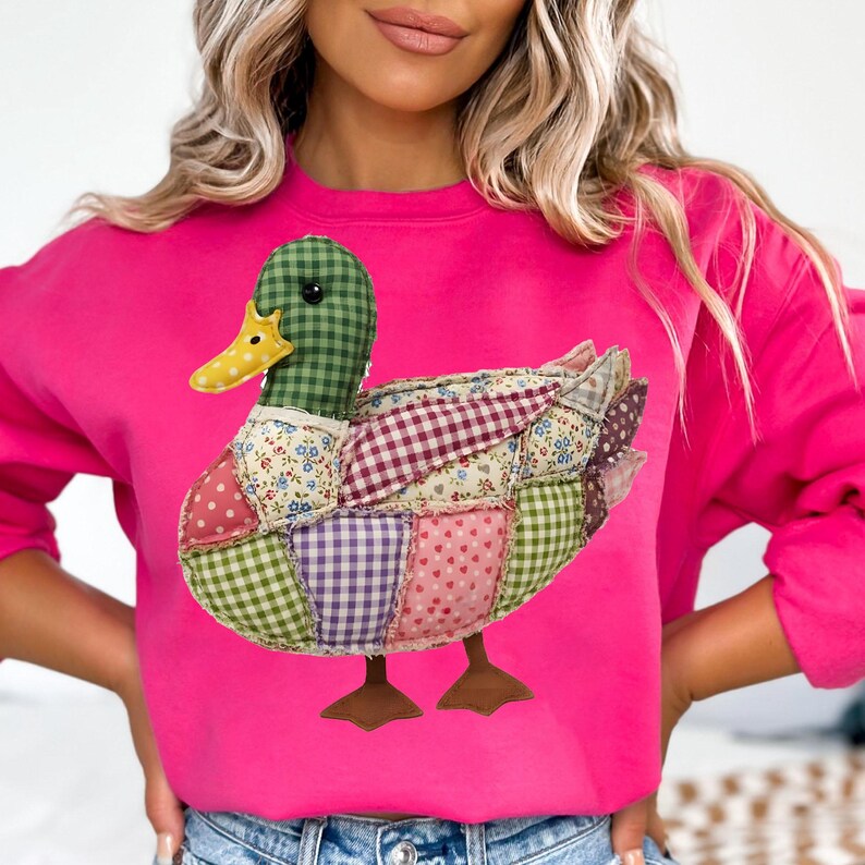 May include: A hot pink sweatshirt featuring a patchwork duck design. The duck has a green checkered head, a yellow beak, and a body made of various patterned fabrics, including floral, polka dot, and gingham. The duck's feet are brown.