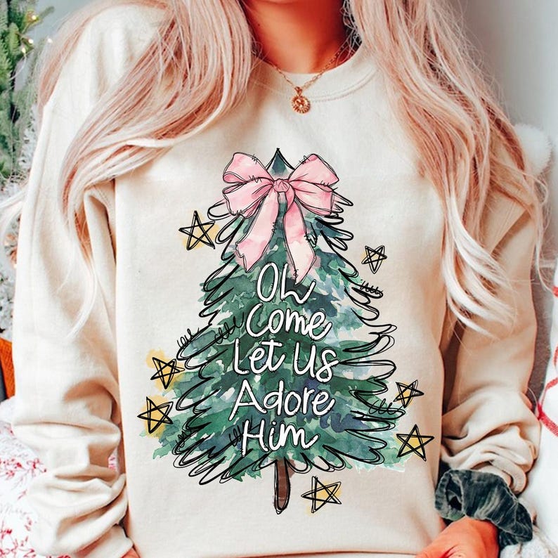 May include: A white sweatshirt with a green watercolor Christmas tree design. The tree has a pink bow and the words "Oh Come Let Us Adore Him" written on it.