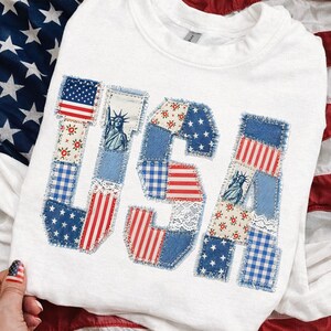 K&ouml;nnte beinhalten: Wei&szlig;es Sweatshirt mit dem Wort "USA" in einem Patchwork-Design appliziert. Die Buchstaben zeigen Denim-, Blumen- und Streifenstoffe sowie Bilder der amerikanischen Flagge und der Freiheitsstatue. Das Sweatshirt wird auf einer Oberfl&auml;che mit einer amerikanischen Flagge pr&auml;sentiert.