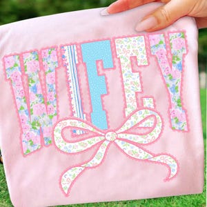 May include: A light pink t-shirt with the word "WIFEY" in a decorative font. The letters are adorned with floral patterns, blue polka dots, and a lace trim. A matching bow is at the bottom. The shirt is held by a hand.