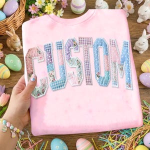 May include: A light pink sweatshirt with the word "CUSTOM" appliqued across the front. The letters are made of patchwork fabric in various pastel patterns. Easter eggs, bunnies, and flowers surround the sweatshirt.