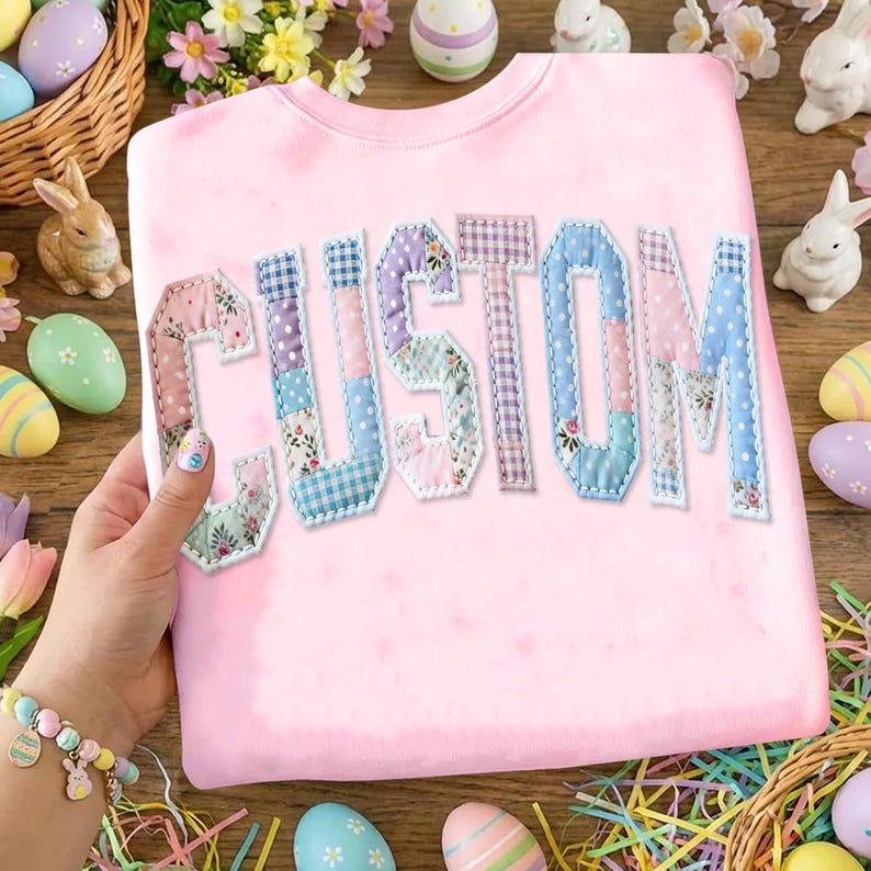 May include: A light pink sweatshirt with the word "CUSTOM" appliqued across the front. The letters are made of patchwork fabric in various pastel patterns. Easter eggs, bunnies, and flowers surround the sweatshirt.