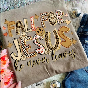 May include: A tan t-shirt with the words "FALL FOR JESUS he never leaves" in a colorful, decorative font. The letters have various patterns, including leopard print, cherries, and checkerboard. A smiley face and floral accents are also included.
