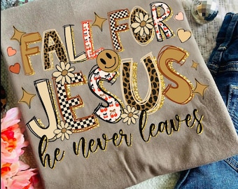 May include: A tan t-shirt with the words "FALL FOR JESUS he never leaves" in a colorful, decorative font. The letters have various patterns, including leopard print, cherries, and checkerboard. A smiley face and floral accents are also included.