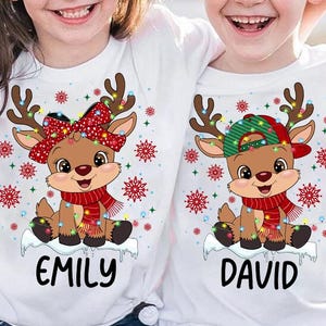 May include: Two white t-shirts with a Christmas reindeer design. One shirt has a reindeer wearing a red bow and the other has a reindeer wearing a green and white hat. The shirts are personalized with the names "Emily" and "David".