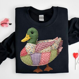 May include: A black sweatshirt featuring a patchwork duck design. The duck has a green checkered head, a yellow beak, and a body composed of various fabric patterns, including polka dots, floral prints, and gingham. The duck has brown feet.