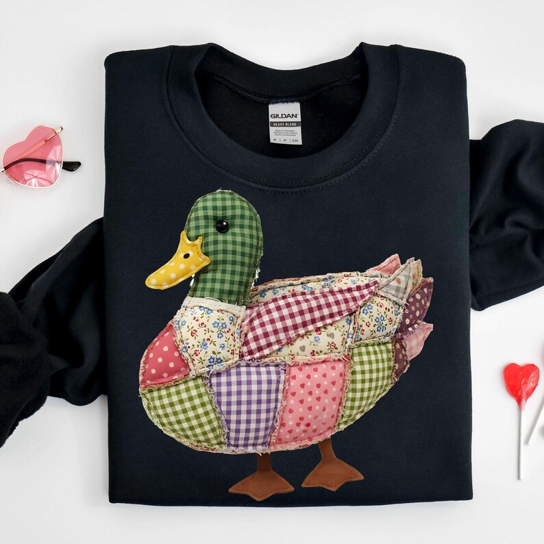 May include: A black sweatshirt featuring a patchwork duck design. The duck has a green checkered head, a yellow beak, and a body composed of various fabric patterns, including polka dots, floral prints, and gingham. The duck has brown feet.