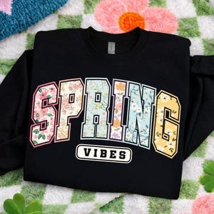 Puede incluir: Sudadera negra con la frase "SPRING VIBES". La palabra "SPRING" tiene un dise&ntilde;o floral y una fuente de estilo universitario. "VIBES" est&aacute; en texto blanco sobre un fondo negro. La sudadera est&aacute; doblada.
