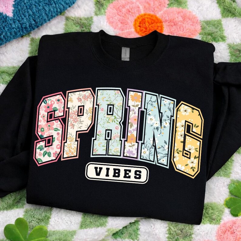 Puede incluir: Sudadera negra con la frase "SPRING VIBES". La palabra "SPRING" tiene un dise&ntilde;o floral y una fuente de estilo universitario. "VIBES" est&aacute; en texto blanco sobre un fondo negro. La sudadera est&aacute; doblada.