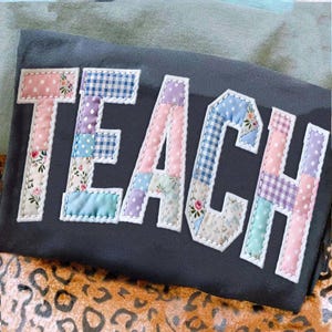 May include: A dark gray fabric item with the word "TEACH" appliqued in colorful patchwork letters. Each letter is composed of various patterned fabrics, including floral, polka dot, and gingham, with white stitching.