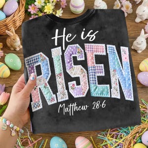 Puede incluir: Sudadera negra con las palabras "He is RISEN" en un dise&ntilde;o de patchwork. El texto "Matthew 28:6" est&aacute; debajo. Decoraciones de Pascua, incluyendo huevos y conejitos, rodean la sudadera.