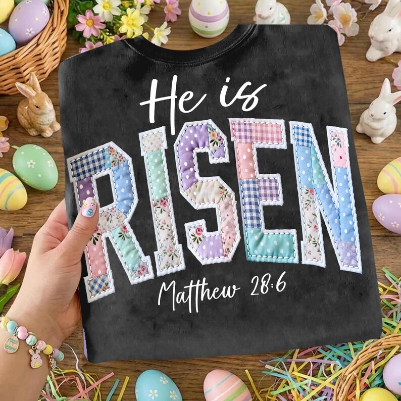Puede incluir: Sudadera negra con las palabras "He is RISEN" en un dise&ntilde;o de patchwork. El texto "Matthew 28:6" est&aacute; debajo. Decoraciones de Pascua, incluyendo huevos y conejitos, rodean la sudadera.