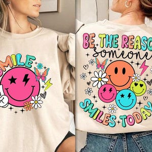 以下が含まれることがあります： ベージュのトレーナーにピンクのスマイリーとカラフルな文字で「SMILE」と書かれています。トレーナーの背面には「BE THE REASON SOMEONE SMILES TODAY」という虹色の文字が書かれています。