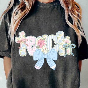 Puede incluir: Una camiseta gris con la palabra "Teach" en letras de patchwork de colores con un lazo azul y detalles florales.