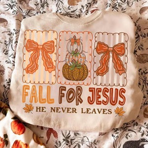 Puede incluir: Sudadera beige con un diseño de temática otoñal. El diseño incluye tres secciones enmarcadas con lazos naranjas y calabazas apiladas. El texto "FALL FOR JESUS" y "HE NEVER LEAVES" está en marrón y naranja.
