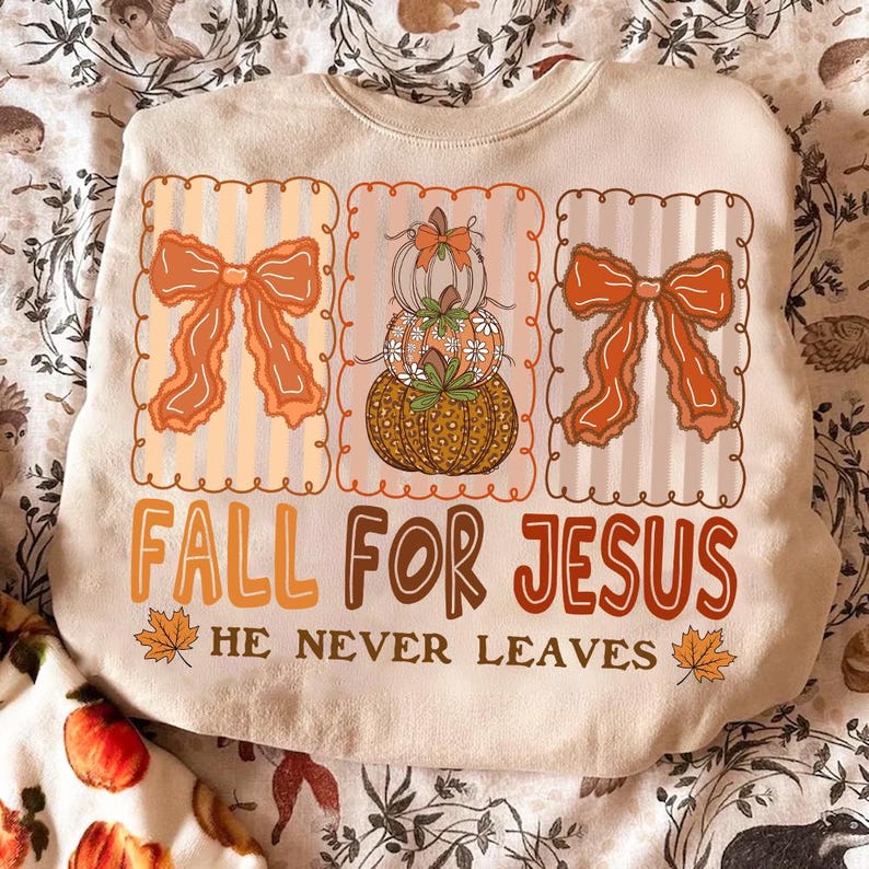 May include: Cream-coloured sweatshirt featuring a seasonal design. The design includes three framed sections with orange bows and stacked pumpkins. The text "FALL FOR JESUS" and "HE NEVER LEAVES" are in brown and orange.