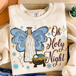 May include: A cream-colored sweatshirt with a Christmas-themed design. The design features an angel with blue wings, a manger with a baby inside, and the text "Oh Holy Night".