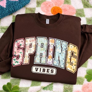 Puede incluir: Una sudadera marr&oacute;n con la palabra "SPRING" en un dise&ntilde;o floral y la palabra "VIBES" en un rect&aacute;ngulo blanco. La sudadera est&aacute; doblada sobre una manta con estampado floral.