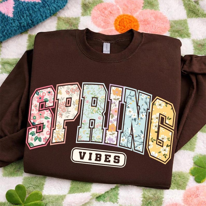 Puede incluir: Una sudadera marr&oacute;n con la palabra "SPRING" en un dise&ntilde;o floral y la palabra "VIBES" en un rect&aacute;ngulo blanco. La sudadera est&aacute; doblada sobre una manta con estampado floral.