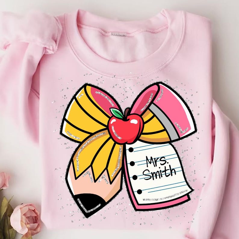 May include: Pink sweatshirt with a glitter design of a yellow and pink bow with a pencil and a notepad that says "Mrs. Smith".