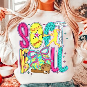 以下が含まれることがあります： ソフトボールの文字と、ソフトボール、バット、グローブ、ボールを打つ選手が描かれたカラフルなグラフィックデザインが施された白いスウェットシャツ。