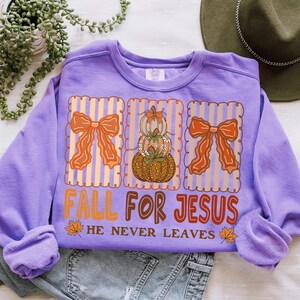 May include: A lilac sweatshirt featuring a fall design. The graphic includes three panels with orange bows and pumpkins. The text "FALL FOR JESUS" is in orange and red, with "HE NEVER LEAVES" below. Autumn leaves are also included in the design.