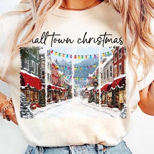 May include: Cream-colored t-shirt featuring a winter scene of a snow-covered town street with shops and colorful lights. The text "Small Town Christmas" is above the image. The shirt is a festive holiday apparel item.
