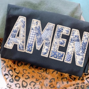 May include: A navy textile featuring the word "AMEN" in appliquéd patchwork letters. The letters are made from blue and white floral and patterned fabrics, stitched with cream thread. The textile rests on a leopard print fabric, with other textiles in the background.