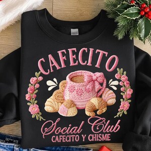 Pode incluir: Camisola preta com design bordado. O design apresenta a palavra "CAFECITO" acima de uma ch&aacute;vena de caf&eacute; rosa com um la&ccedil;o, rodeada por flores e croissants. Abaixo est&aacute; "Social Club" e "CAFECITO Y CHISME".