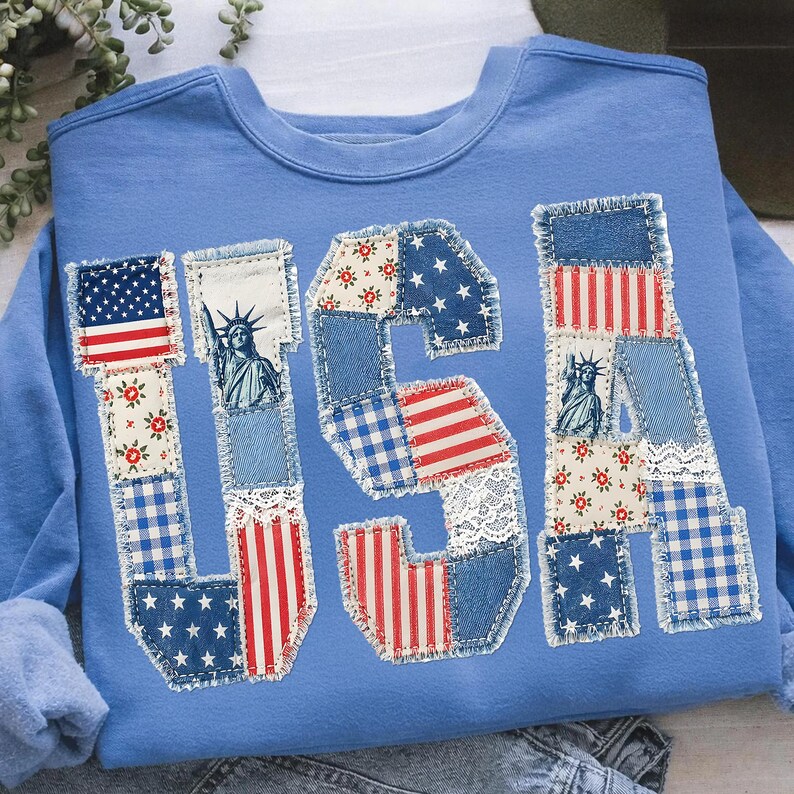 K&ouml;nnte beinhalten: Hellblaues Sweatshirt mit dem Wort "USA" auf der Vorderseite. Die Buchstaben bestehen aus verschiedenen Stoffflicken, darunter die amerikanische Flagge, Blumenmuster und die Freiheitsstatue.