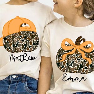 May include: Two cream-colored t-shirts with pumpkin designs. One shirt features a pumpkin wearing an orange baseball cap, with the name "Mathew" below. The other shirt has a pumpkin with an orange bow and the name "Emma".