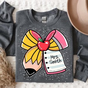 May include: A grey sweatshirt featuring a sparkly pencil bow design. The bow is made of yellow and pink pencils with a red apple in the centre. The text "Mrs. Smith" is written on a pink notepad.