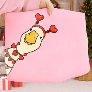 May include: A pink pillow featuring a cartoon duck with a yellow beak and a red heart-shaped headband. The duck is adorned with heart-shaped accents, creating a whimsical and festive design. The pillow is held by a person.