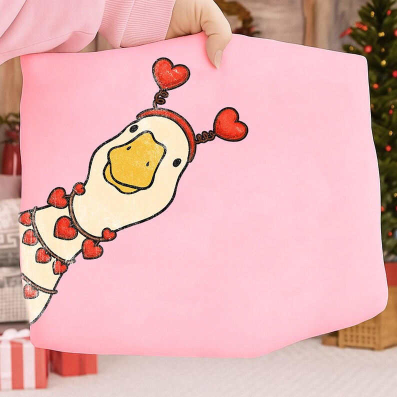 May include: A pink pillow featuring a cartoon duck with a yellow beak and a red heart-shaped headband. The duck is adorned with heart-shaped accents, creating a whimsical and festive design. The pillow is held by a person.