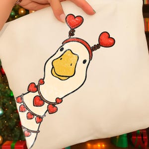 May include: A cream-colored pillow featuring a cartoon goose with a yellow beak and a red heart-shaped headband. The goose has a necklace of red hearts. The pillow is held up by a person.