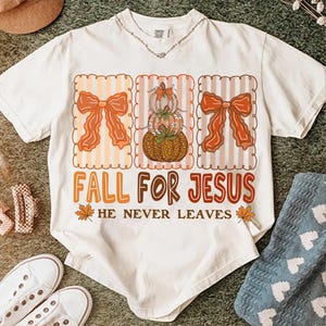 May include: Cream-coloured t-shirt with a fall-themed design. The design includes three decorative frames with orange bows and pumpkins. The text "FALL FOR JESUS" is displayed below the frames, with the phrase "HE NEVER LEAVES" underneath. The shirt is surrounded by other items.