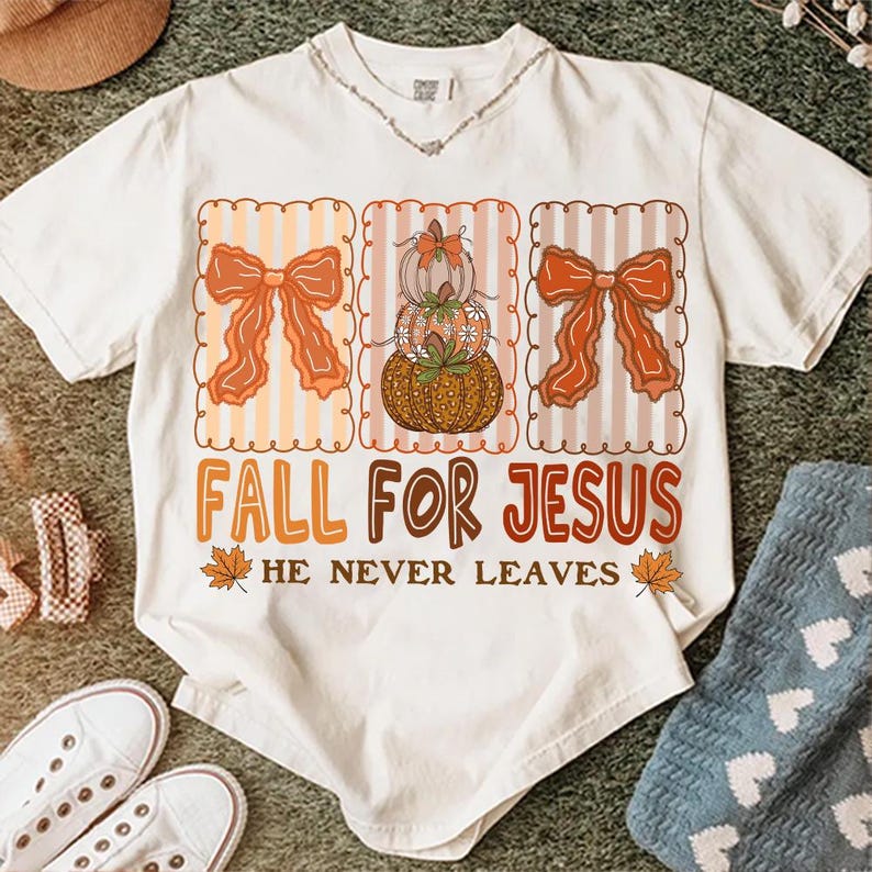 May include: Cream-coloured t-shirt with a fall-themed design. The design includes three decorative frames with orange bows and pumpkins. The text "FALL FOR JESUS" is displayed below the frames, with the phrase "HE NEVER LEAVES" underneath. The shirt is surrounded by other items.