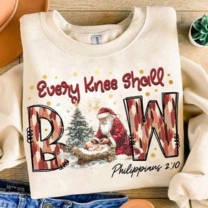May include: A cream-colored sweatshirt with a Christmas-themed design. The design features the words "Every Knee Shall Bow" with the letters "B" and "W" stylized in a red and white pattern. A depiction of Santa Claus kneeling before the baby Jesus in a manger is in the center of the design. The text "Philippians 2:10" is at the bottom of the design.