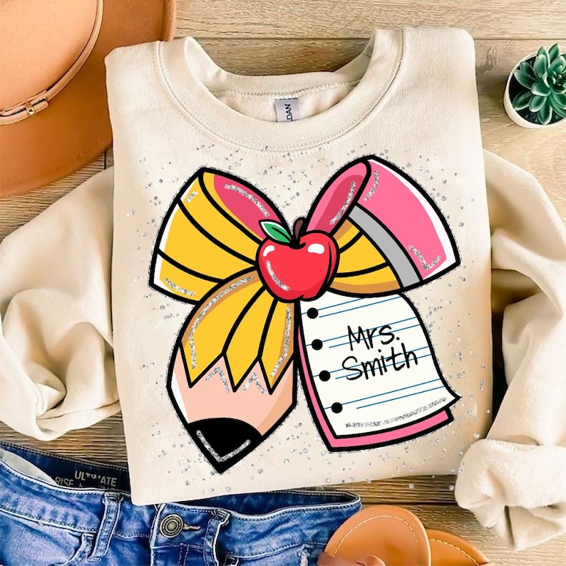 May include: A beige sweatshirt with a glitter design of a pencil bow with a red apple and a notepad that says "Mrs. Smith".