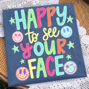K&ouml;nnte beinhalten: Ein dunkelblaues T-Shirt mit dem bunten Schriftzug HAPPY to see YOUR FACE in Rosa-, Orange-, Gelb- und Hellgr&uuml;nt&ouml;nen. Das Design enth&auml;lt vier Smiley-Gesichter mit verschiedenen Mustern sowie mehrere kleine hellgr&uuml;ne Sterne.