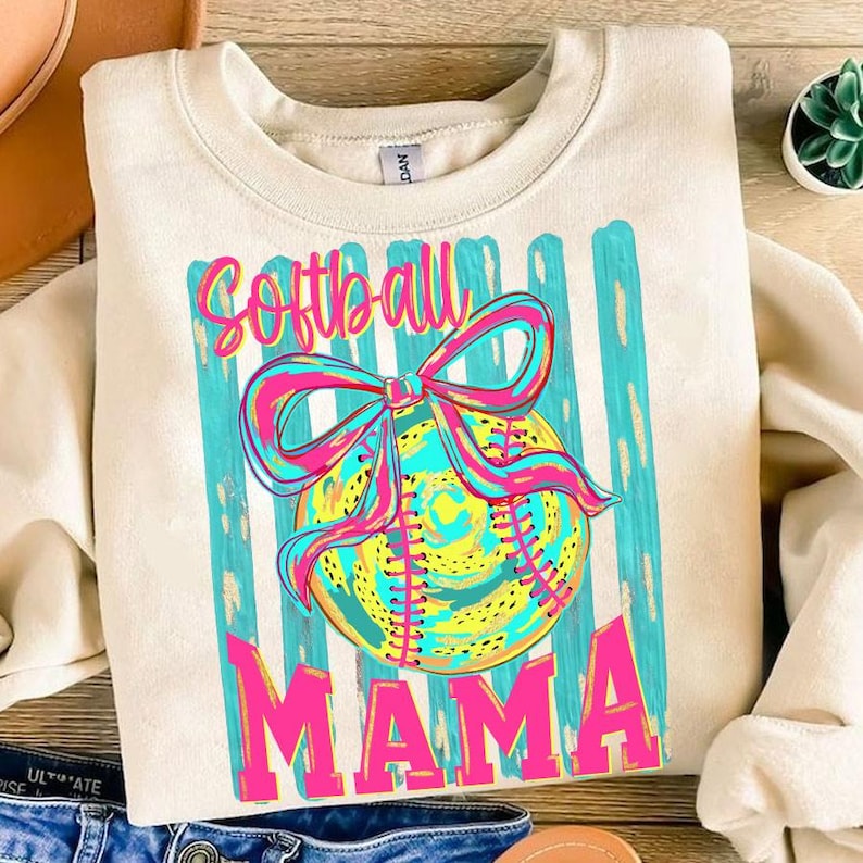 May include: Cream-colored sweatshirt with a colorful softball graphic. The design features a softball with a bow, teal stripes, and the words "Softball Mama" in pink and teal. A casual, sporty top.