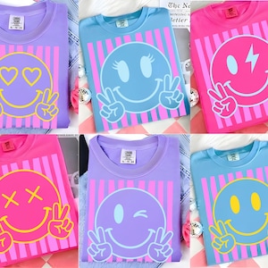 Könnte beinhalten: Eine Auswahl an farbenfrohen T-Shirts mit Smiley-Gesichtern. Jedes Shirt hat einen anderen Smiley-Gesichtsausdruck, darunter Herzen, Zwinker und Friedenszeichen. Die Shirts sind in verschiedenen Farben erhältlich, darunter Lila, Pink und Blau, mit vertikal gestreiften Hintergründen.