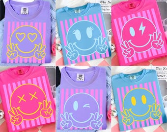 May include: A collection of colorful t-shirts featuring smiley face designs. Each shirt has a different smiley face expression, including hearts, winks, and peace signs. The shirts are in various colors, including purple, pink, and blue, with vertical striped backgrounds.