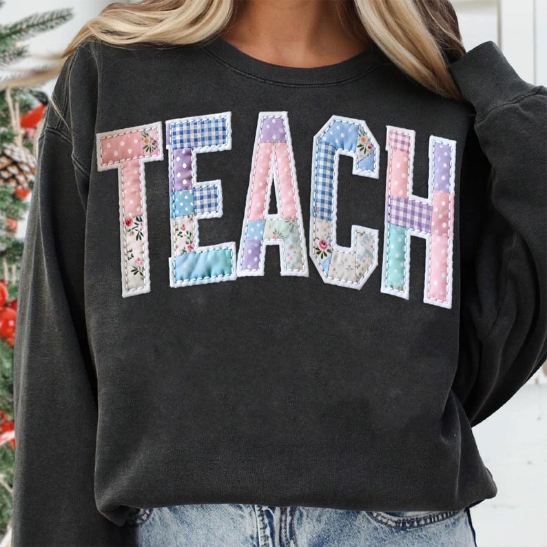 Może przedstawiać: Ciemnoszara bluza z napisem "TEACH" w dużych, patchworkowych literach na piersi. Litery wykonane są z r&oacute;żnych tkanin w pastelowych kolorach, w tym wzor&oacute;w kwiatowych i w kratę. Bluza ma okrągły dekolt i długie rękawy.
