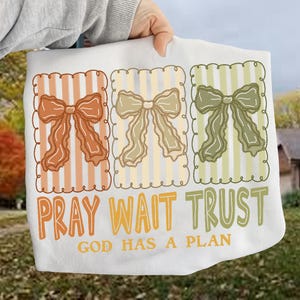 May include: White decorative towel featuring three rectangular panels with bow designs in orange, beige, and green. The towel has the words "PRAY WAIT TRUST" and "GOD HAS A PLAN" printed in orange and green.