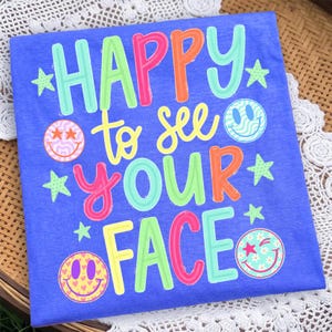 K&ouml;nnte beinhalten: Ein blaues T-Shirt mit dem mehrfarbigen Text HAPPY to see YOUR FACE in verschiedenen Schriftarten und Farben. Das Design enth&auml;lt vier verschiedene Smiley-Gesichter mit Mustern wie Sternen, Wellen und Herzen sowie mehrere kleine gr&uuml;ne Sterne.