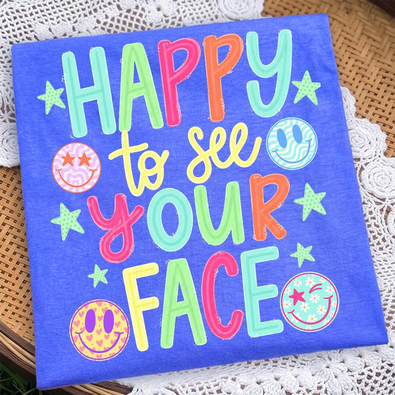 K&ouml;nnte beinhalten: Ein blaues T-Shirt mit dem mehrfarbigen Text HAPPY to see YOUR FACE in verschiedenen Schriftarten und Farben. Das Design enth&auml;lt vier verschiedene Smiley-Gesichter mit Mustern wie Sternen, Wellen und Herzen sowie mehrere kleine gr&uuml;ne Sterne.