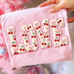 May include: A light pink shirt with the word "LOVE" embroidered in a white, textured font. The letters are decorated with red cherries, pink bows, and small pink hearts. The shirt is held up by a hand.